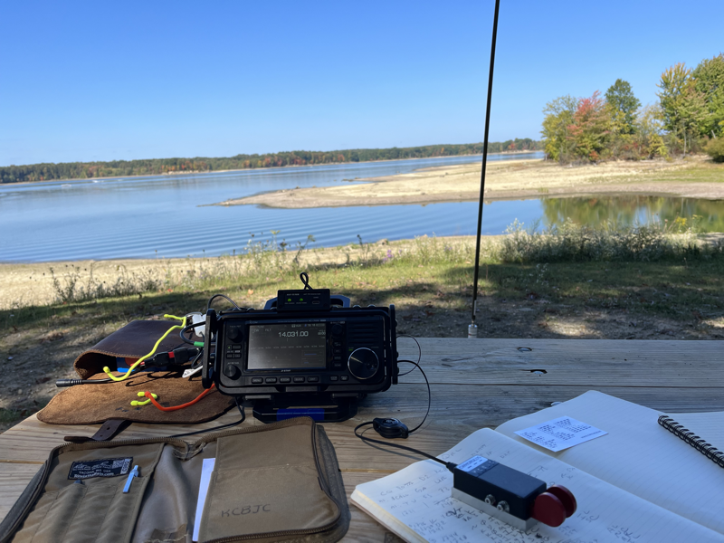 IC-705, notebook, and paddles on a picnic table with a view of the water. A Chameleon vertical antenna is in the background.