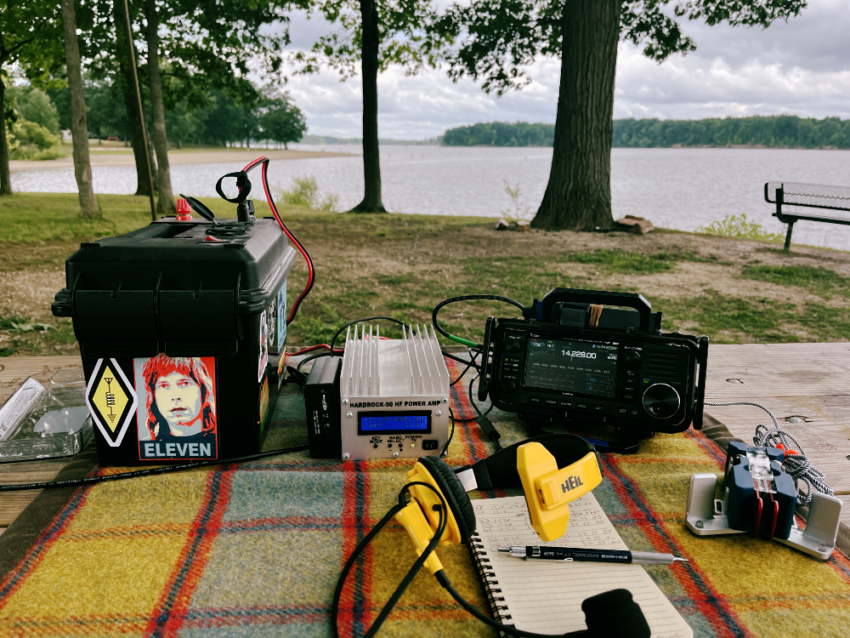 Battery Box, HR50, IC-705, Paddles, Headset, and log book with pencil on a tarp on a picnic table. There are trees and water in the background.