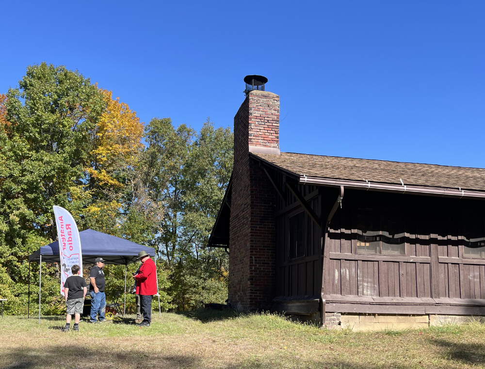 Club President, Assistant Cubmaster, and Cub Scout standing near a popup shelter and next to a cabin with the PCARS banner flying proudly.