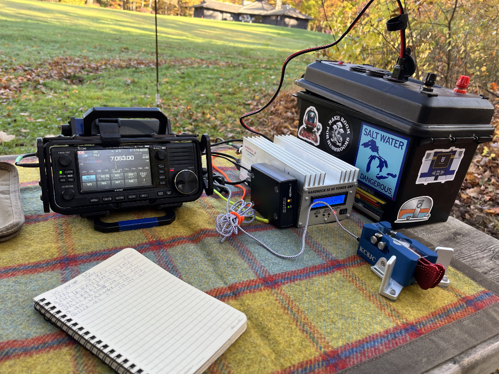 IC-705, Hardrock-50, battery box, and Begali Paddles next to a log book on a plaid tarp resting on a picnic table.