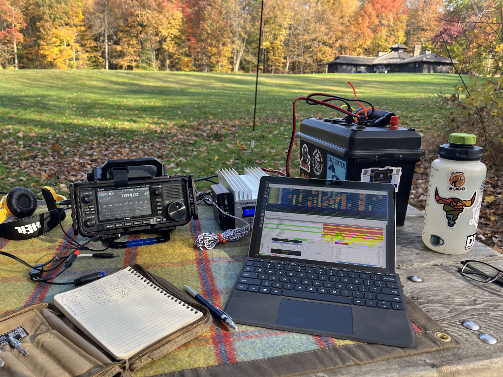 IC-705, Hardrock-50, battery box, and Surface tablet next to a log book on a plaid tarp resting on a picnic table.