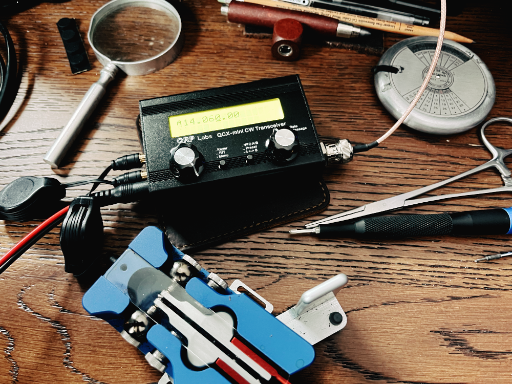 The completed and powered up QCX-mini on a desk next to a magnifying glass, Begali Traveller paddles, a hemostat, and a screwdriver.