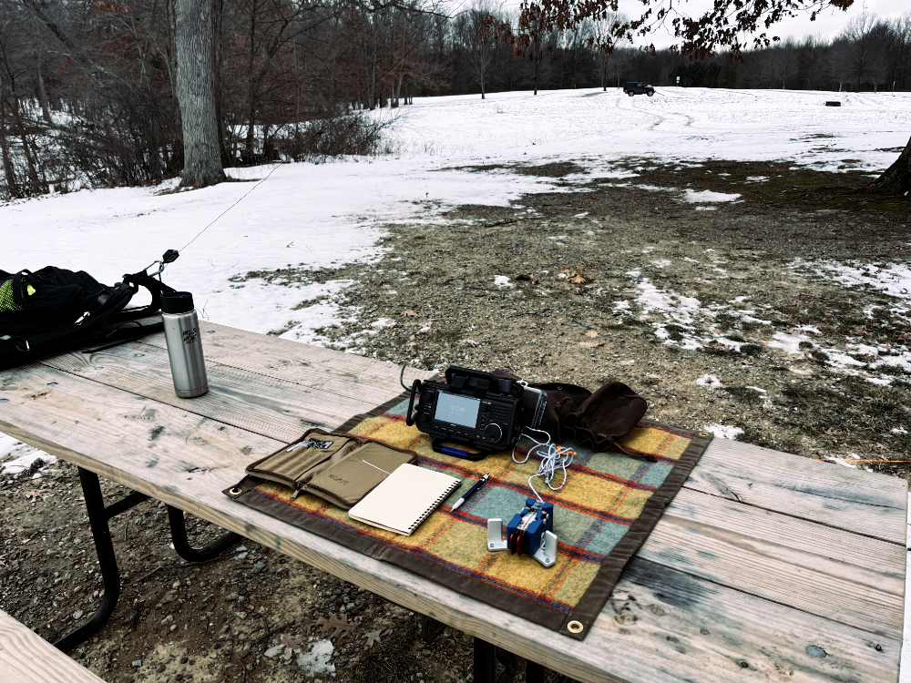 A picnic table with a backpack, coffee cup, IC-705, log book, paddles, pencil and assorted bags on a plaid tarp.