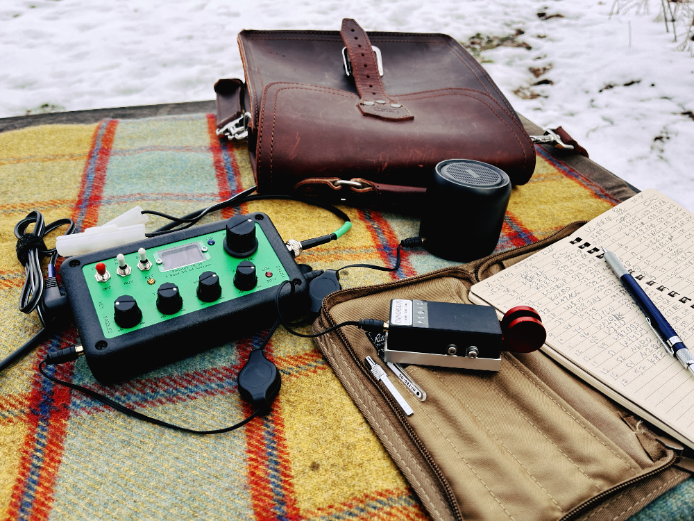 The Penntek TR-35 with a green face and assorted switches and knobs on a plaid tarp next to a leather sachel, CW Morse aluminum paddles, Anker portable speaker, and a log book with pencil. It is all haphazardly strewn about by someone who was in a hurry to set up.